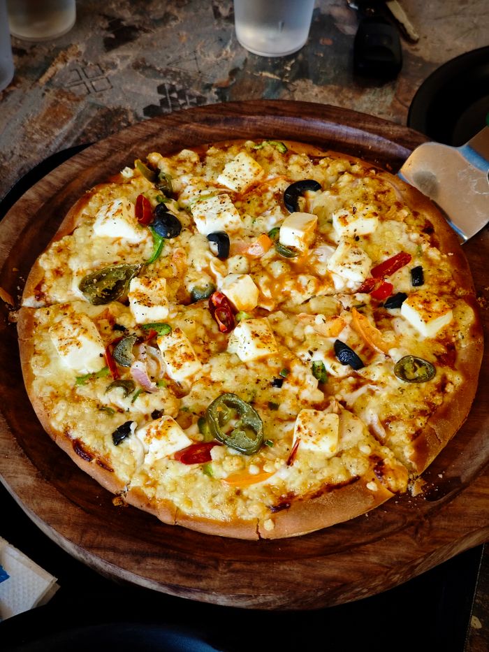 Close-up of a delicious veggie pizza with paneer, olives, jalapenos, and spices in a restaurant setting.