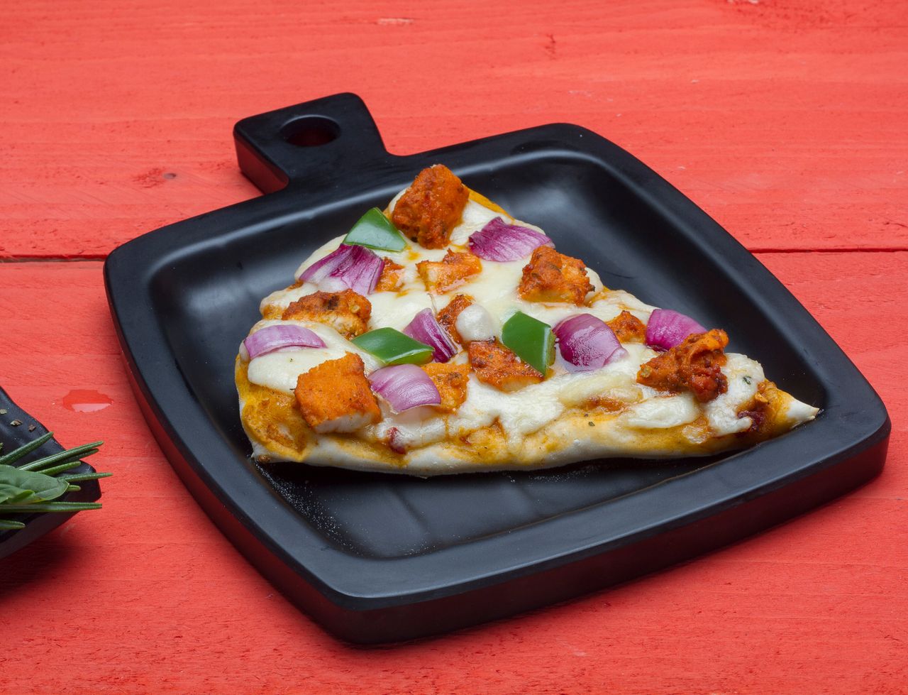 Delicious veggie pizza slice with toppings on a black tray against red background.